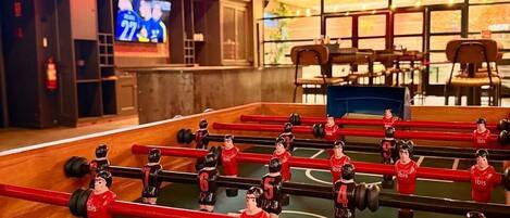 Uma mesa de futebol de mesa em um ambiente com uma placa 'BRAVAR' visível, uma tela de televisão e uma área de estar com cadeiras.