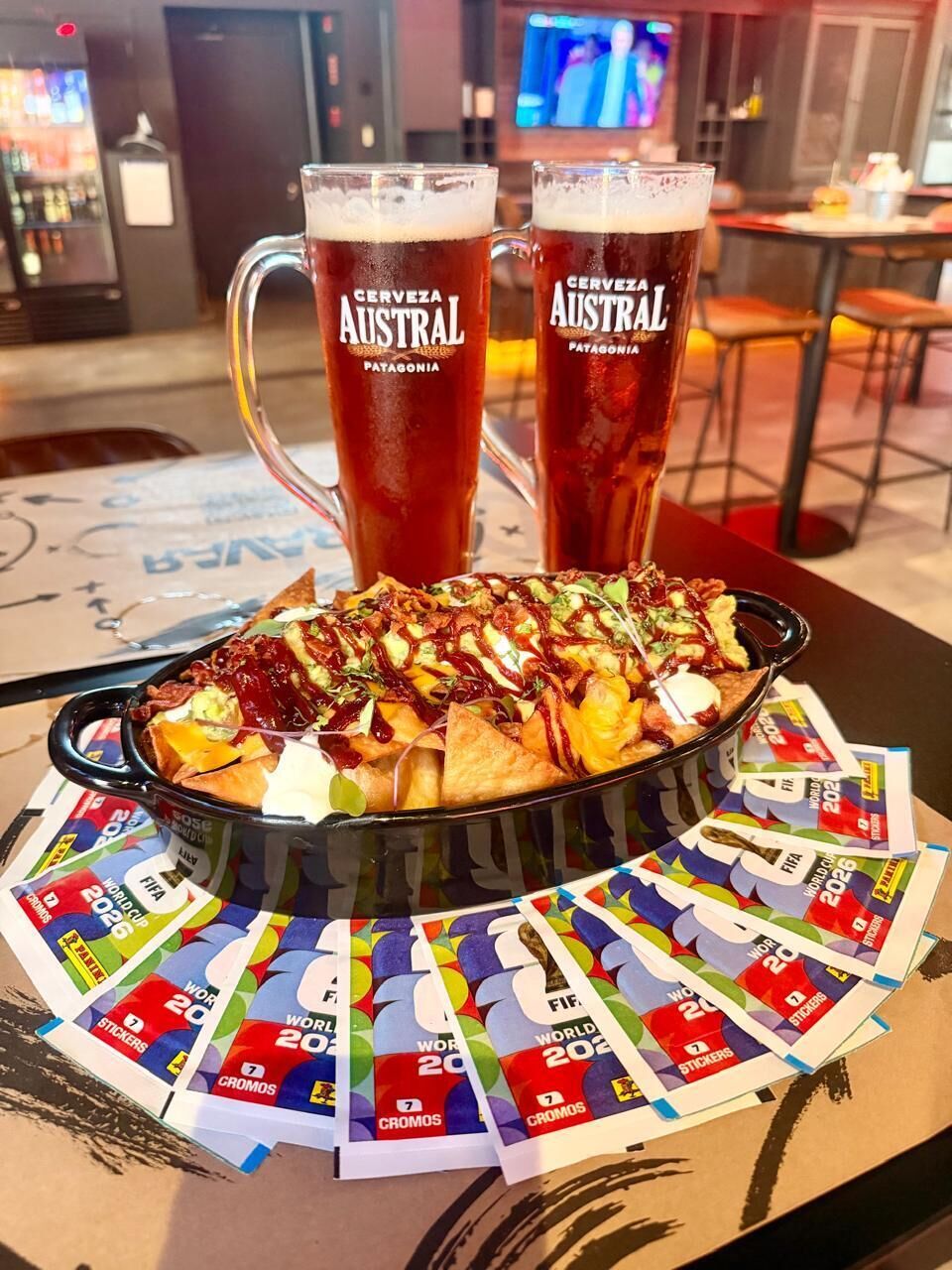 Una comida con nachos y dos jarras de cerveza etiquetadas 'Cerveza Austral Patagonia' sobre una mesa con pegatinas de la Copa Mundial de la FIFA 2022.