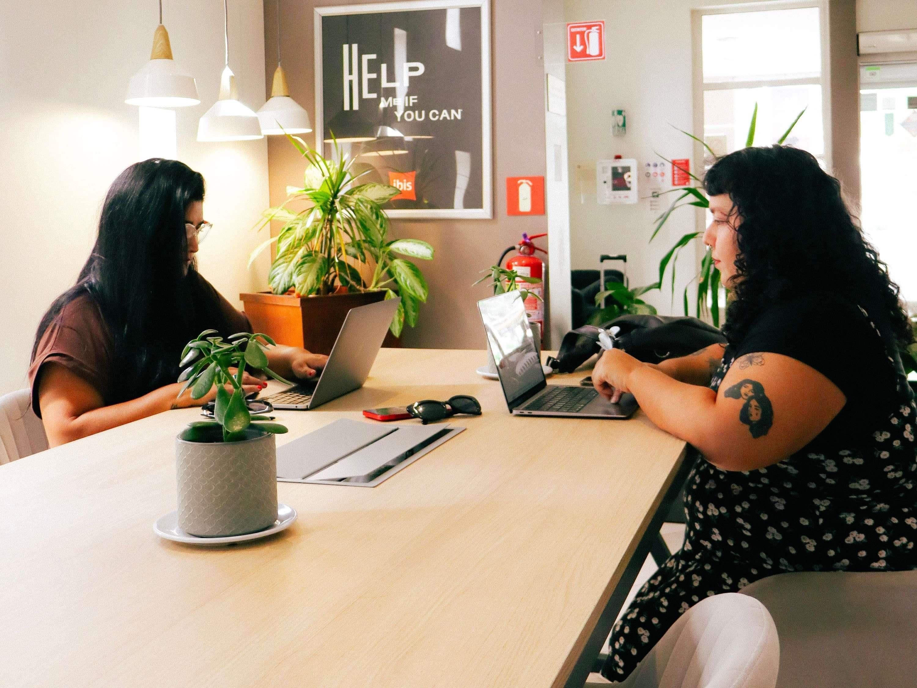Dos personas sentadas en una mesa con computadoras portátiles, rodeadas de plantas en macetas, en una habitación con lámparas colgantes y un letrero de seguridad contra incendios.