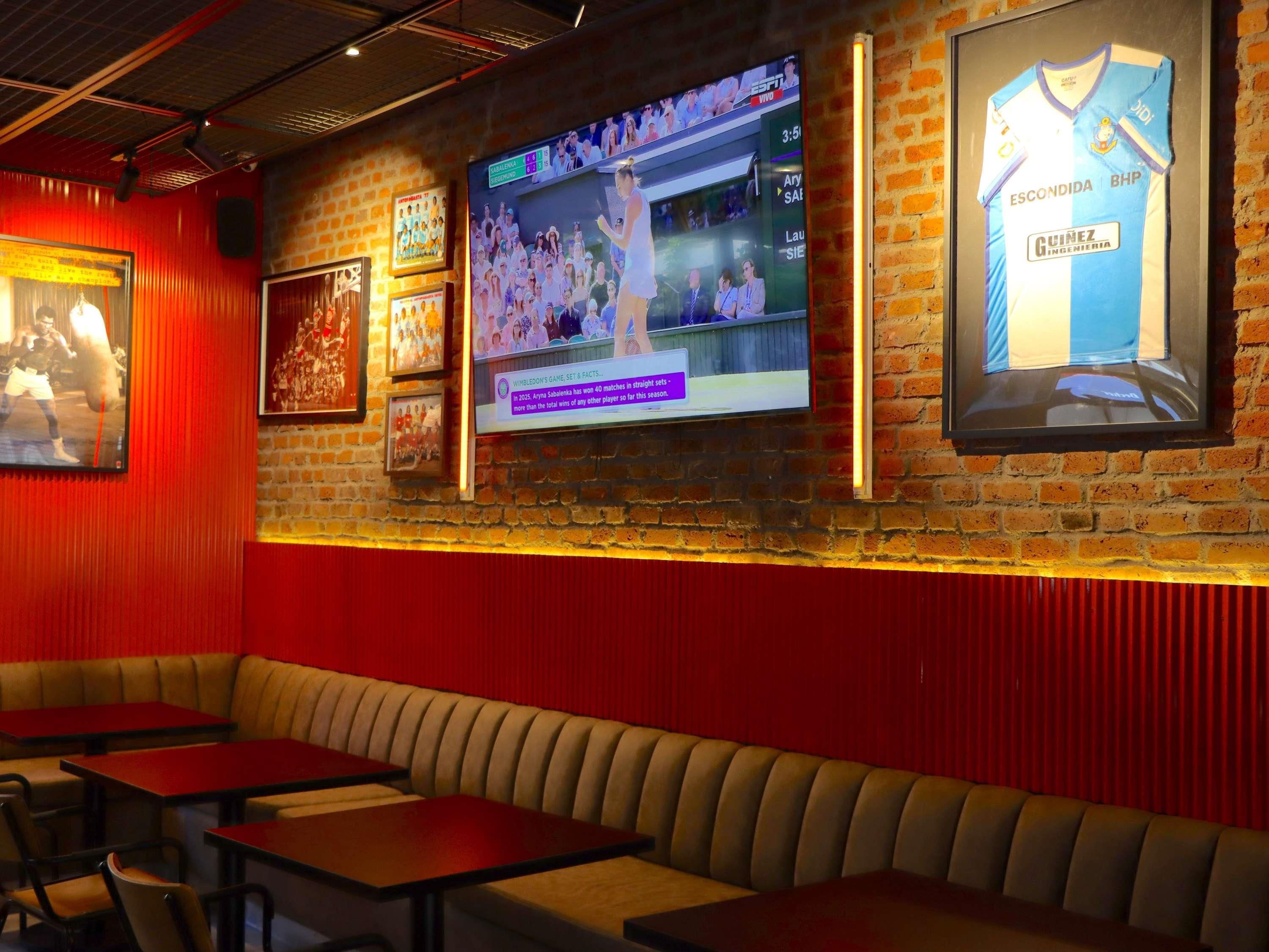 Un bar deportivo con una pantalla grande mostrando un evento deportivo, zonas de estar con mesas y cabinas, y recuerdos deportivos enmarcados en las paredes.