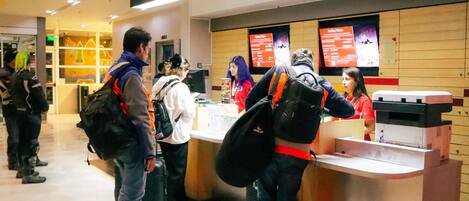 Una fila en un mostrador con personas esperando, una impresora y un panel de menú al fondo.