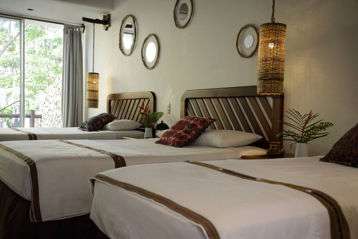 A hotel room with two beds, each with a headboard and pillows, a large window with curtains, and wall-mounted mirrors.