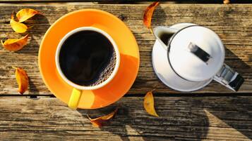 A cup of coffee on an orange saucer, a white teapot, and scattered autumn leaves on a wooden surface.