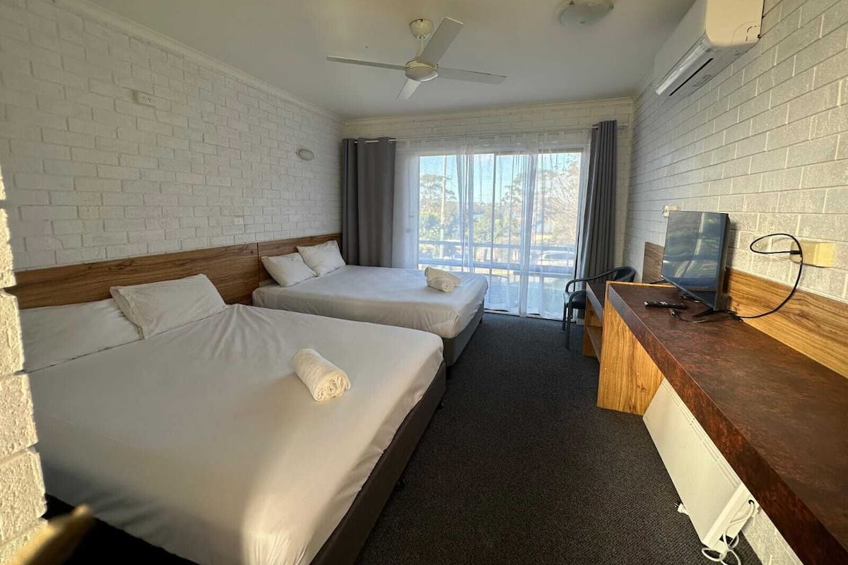 A hotel room with two beds, a desk with a TV, a ceiling fan, and a large window with curtains.