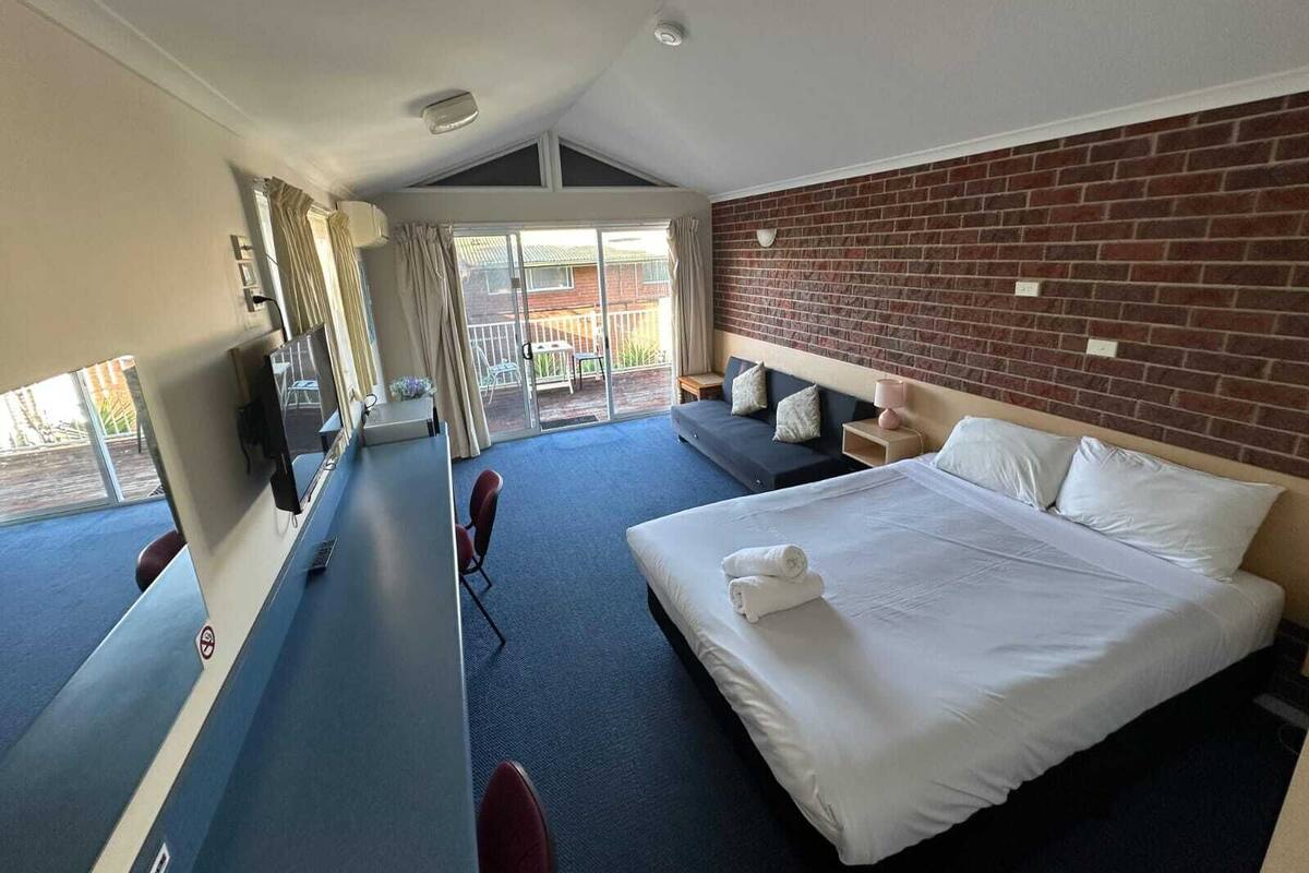 A hotel room with a large bed, a desk, a chair, a TV, and a view of the outdoor patio.