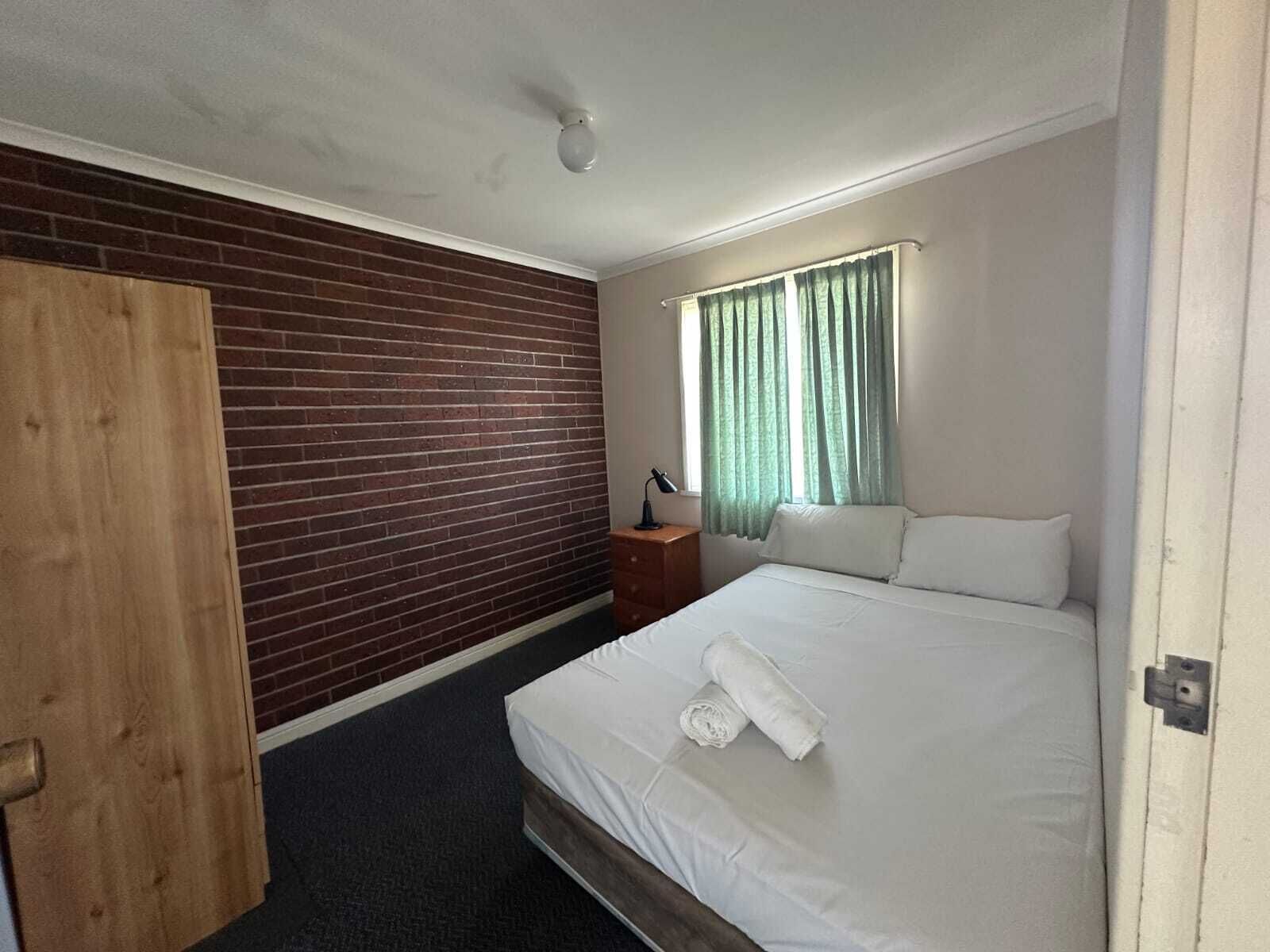 Une chambre d’hôtel avec un mur d’accent en briques, un lit avec du linge de lit blanc, une porte en bois, une table de chevet avec une lampe et une fenêtre avec des rideaux verts.
