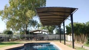 A swimming pool with a covered patio area, surrounded by a fence and trees.