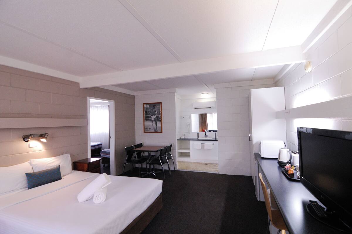 Hotellihuone, jossa on sänky, työpöytä tietokoneella, televisio ja pieni keittiönurkkaus.