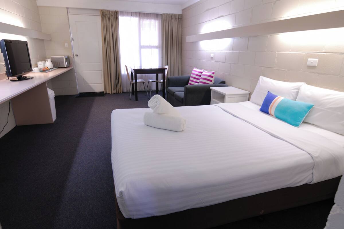 Hotellihuone, jossa on sänky, työpöytä tietokoneella, tuoli ja pieni pöytä.