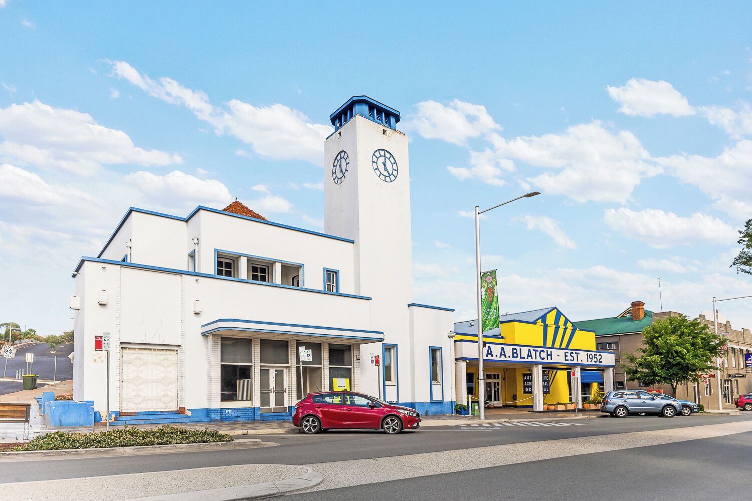 A building with a clock tower, a red car parked in front, and a sign for A.A. Blatch - East 1952.