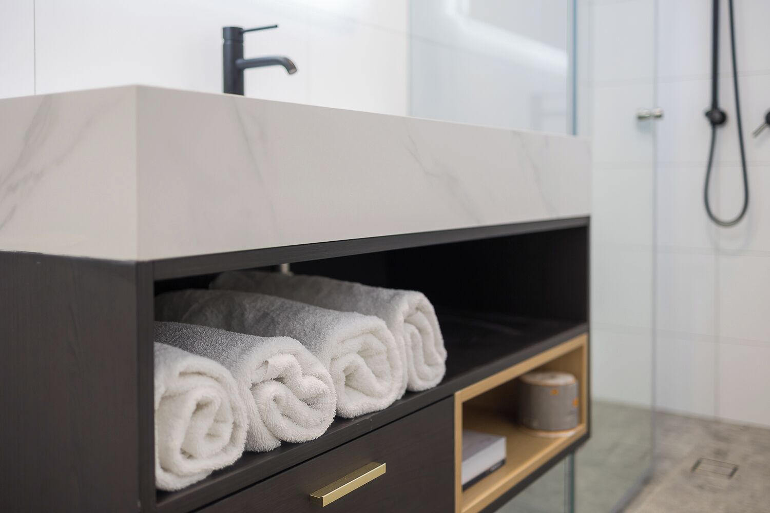 Un tocador de baño con encimera de mármol, un perchero con toallas dobladas y un estante de madera con un rollo de papel higiénico.