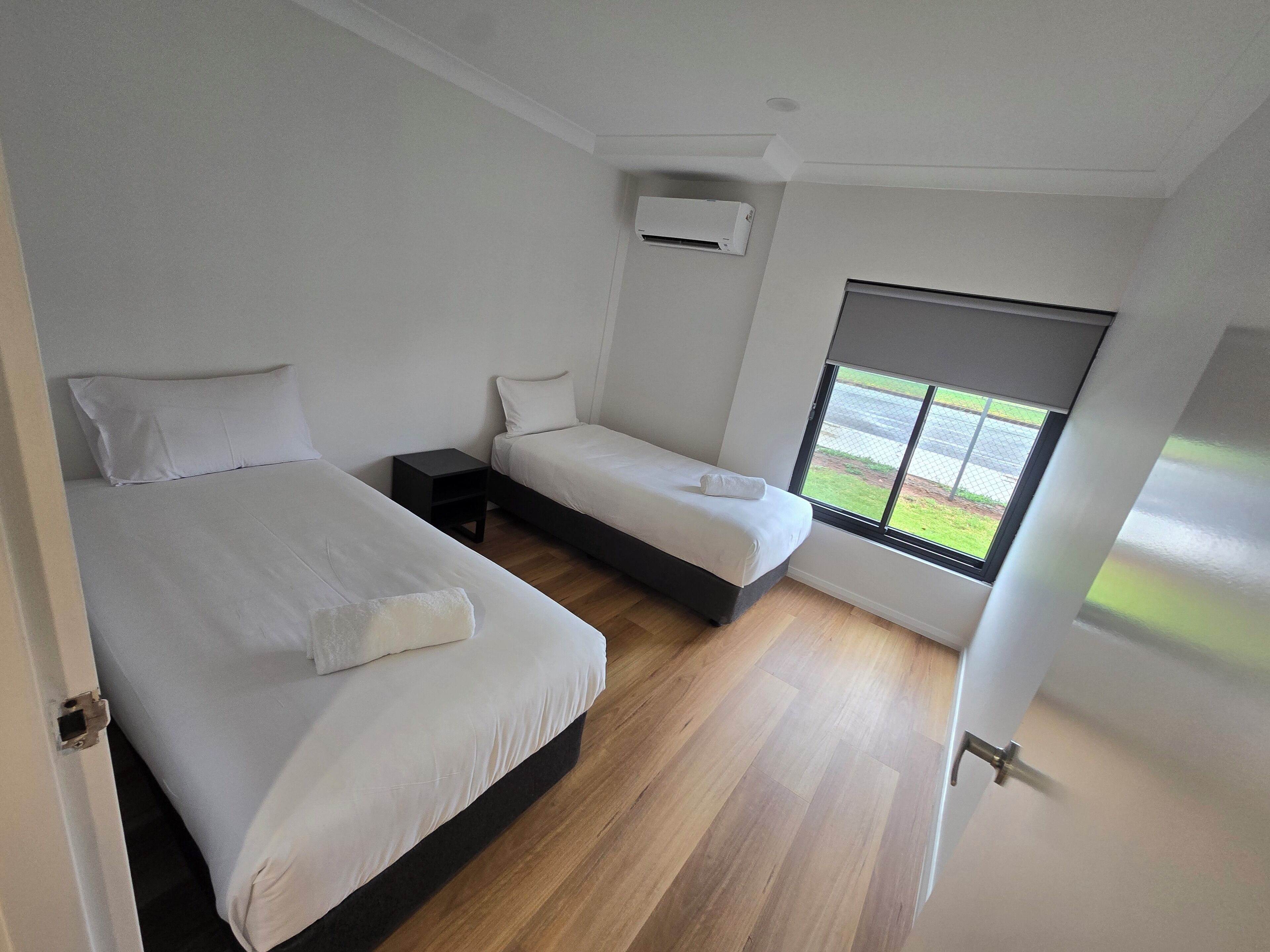 A bedroom with two beds, a small bedside table, a window with a view of the outdoors, and an air conditioning unit on the wall.