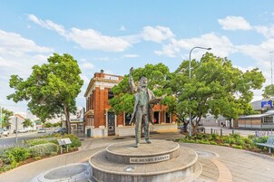 Una estatua de Sir Henry Parkes en una plaza pública, con un edificio de ladrillo al fondo.
