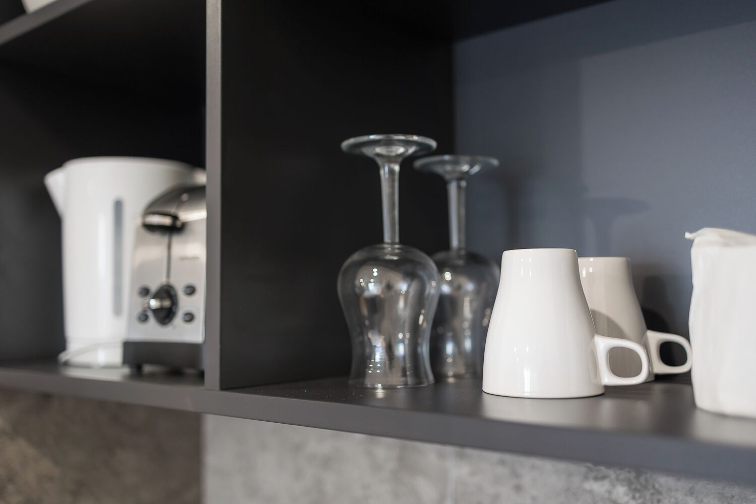 A kitchen shelf with a coffee maker, wine glasses, and white mugs.