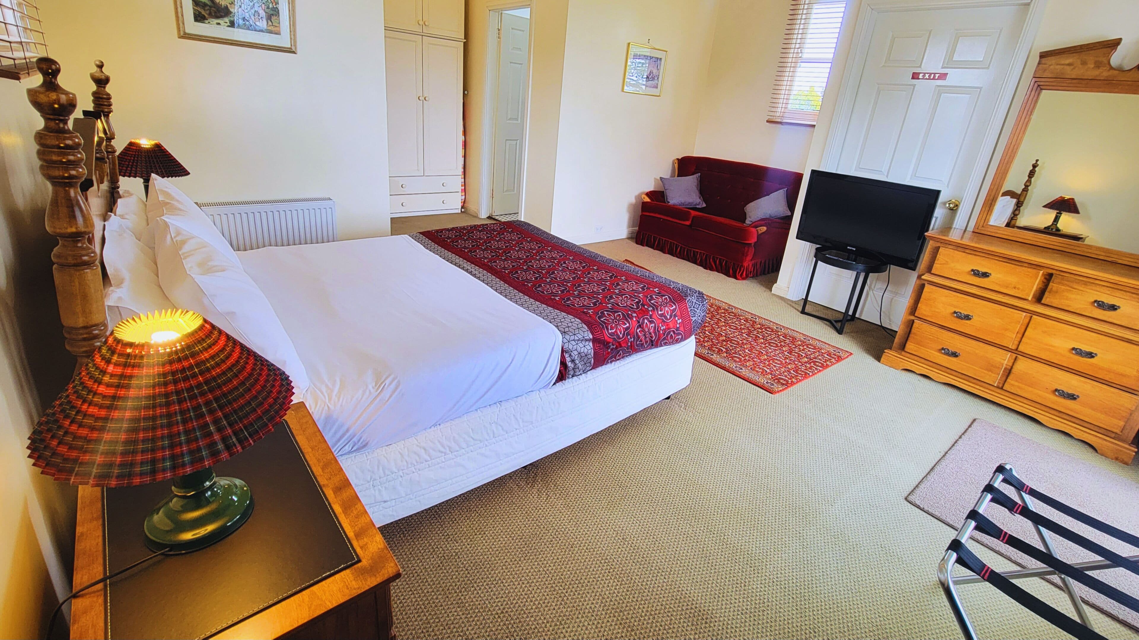 A bedroom with a bed, a desk with a lamp, a television, a dresser, and a red sofa.