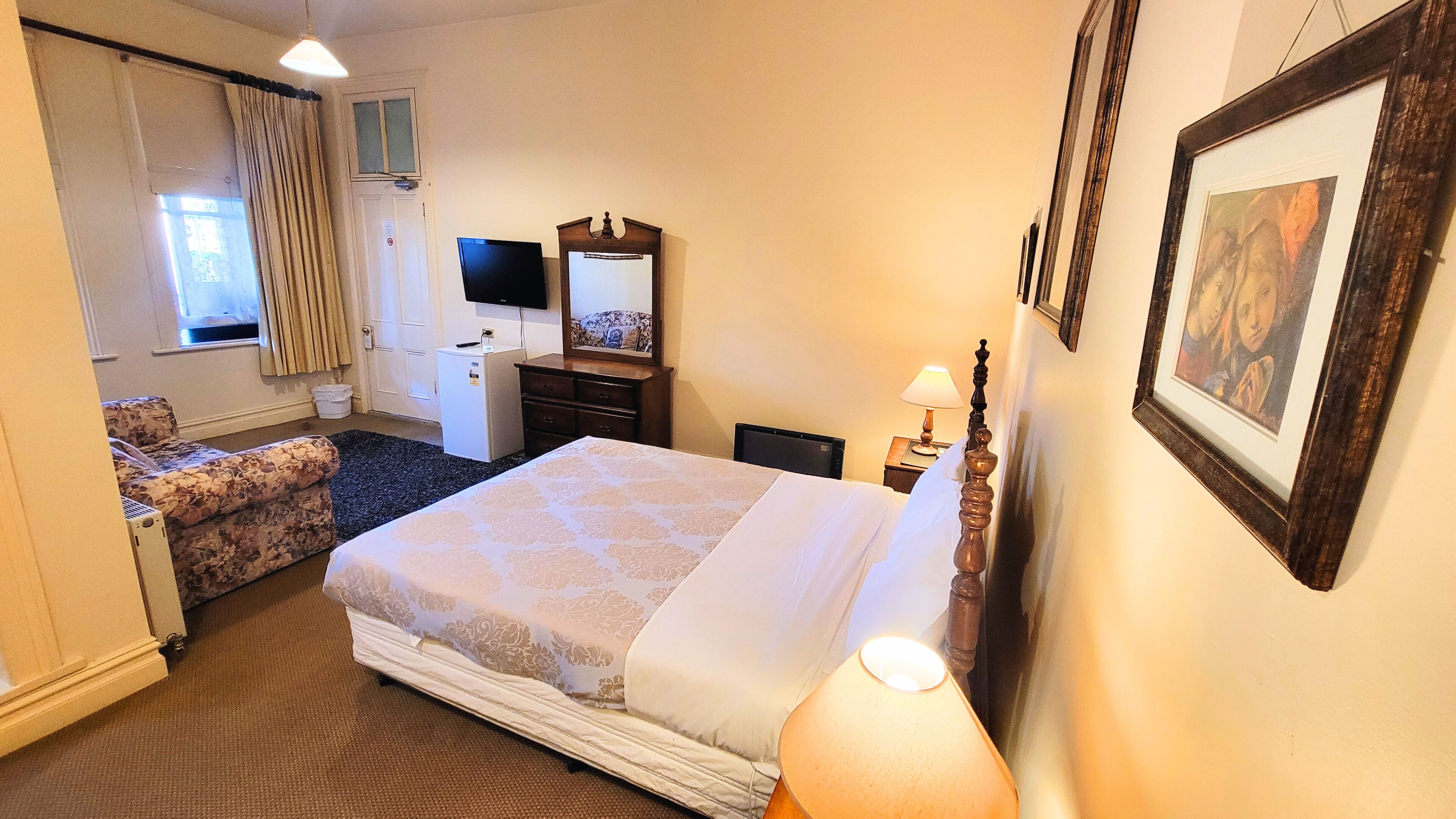 Una habitación de hotel con cama, escritorio, silla, televisor, espejo y un cuadro en la pared.