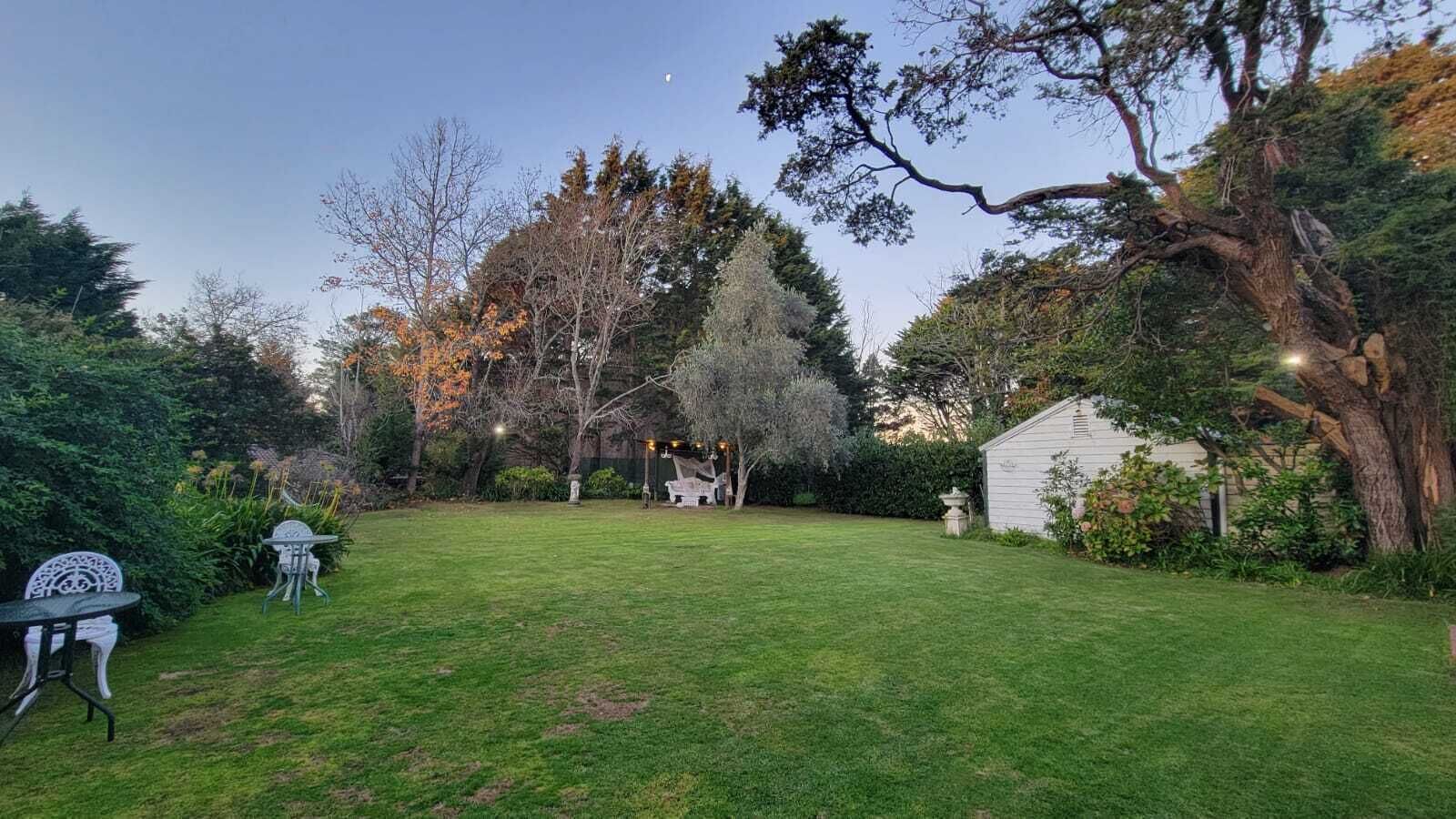 Un patio trasero bien cuidado con césped, una mesa de jardín y sillas, rodeado de árboles y un cobertizo.