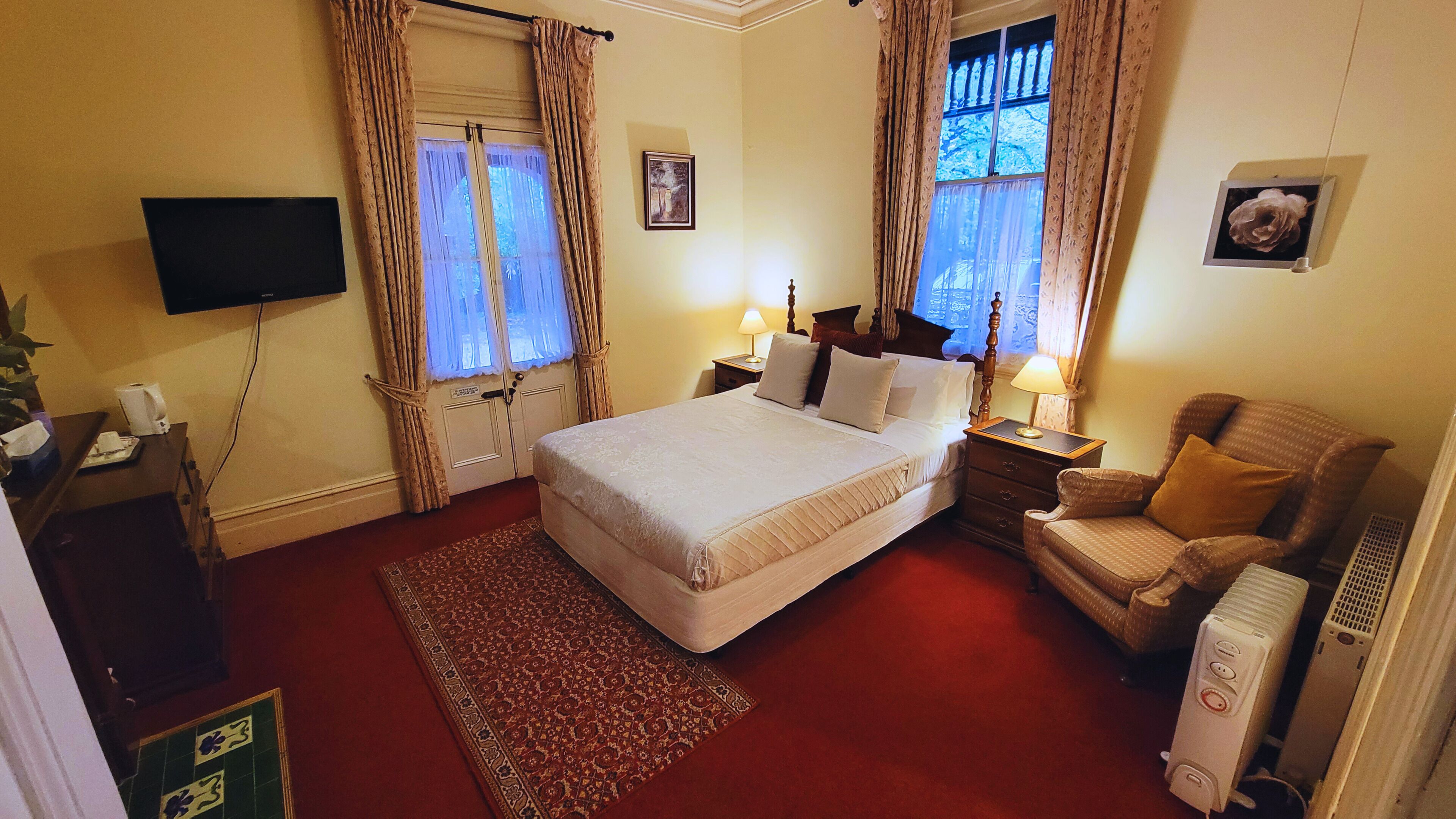 A hotel room with a bed, a chair, a desk with a lamp, a television, and a radiator.