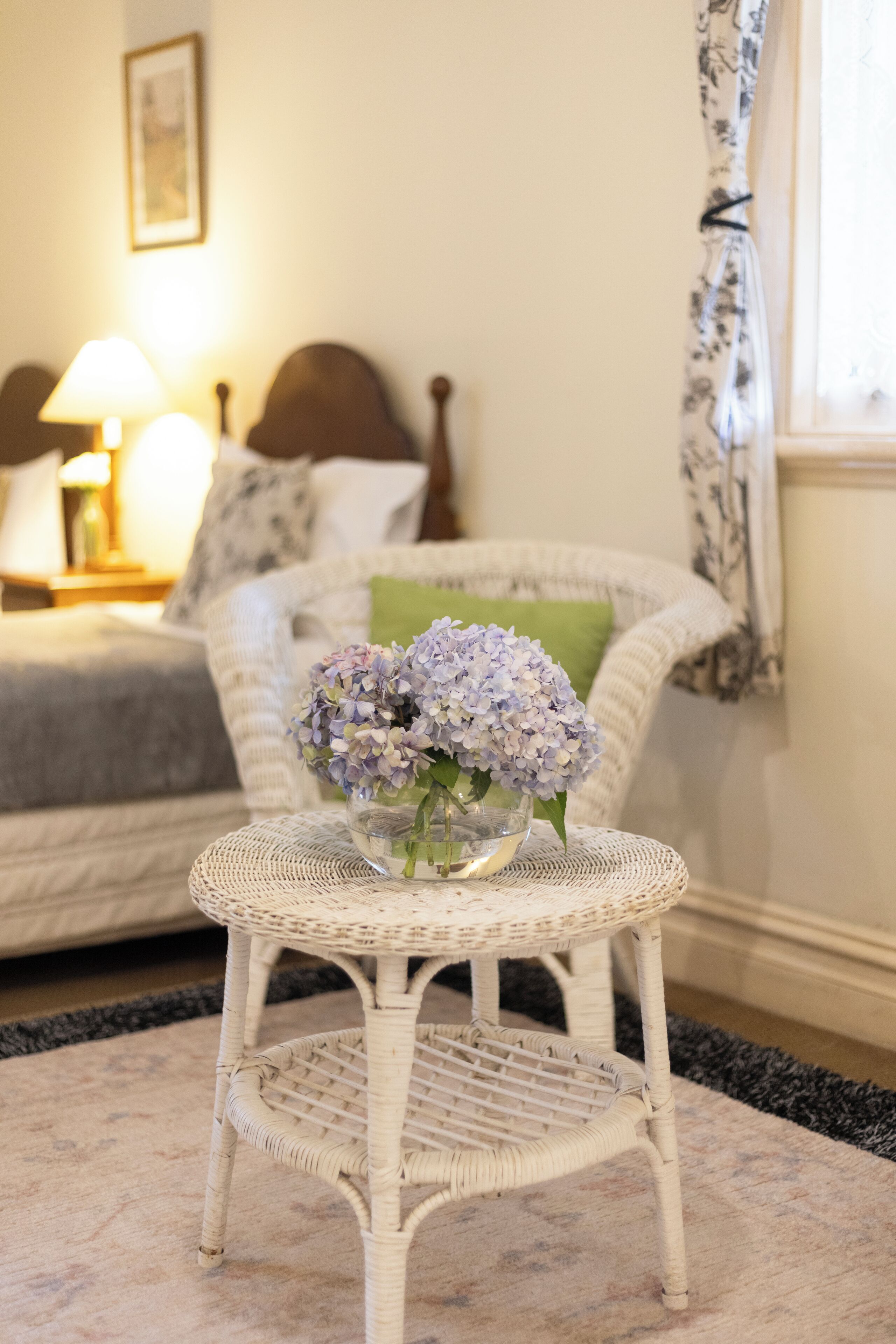 Una silla de mimbre con un jarrón de vidrio con flores, una cama con cabecera, una lámpara y una ventana con cortinas.
