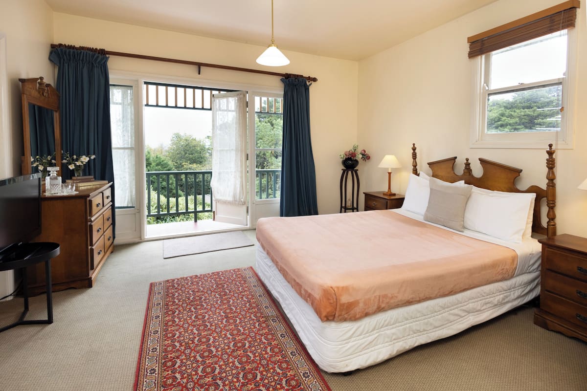 A bedroom with a large bed, a desk, a chair, a dresser, a nightstand, a lamp, and a window with a view of greenery.