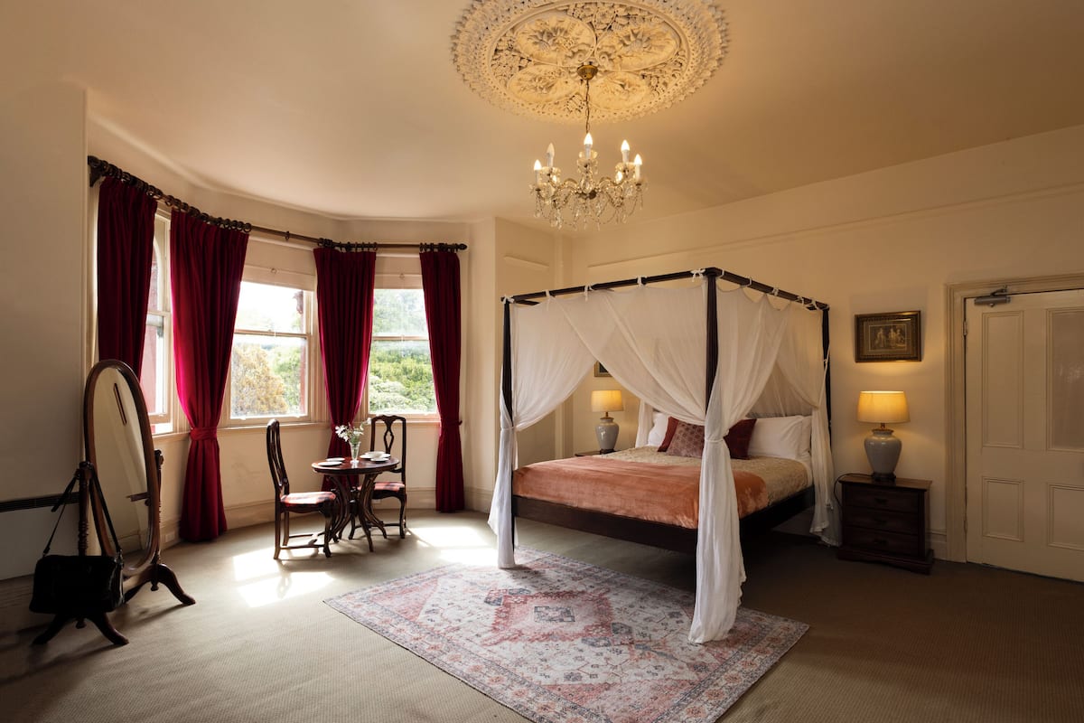 A four-poster bed with a canopy, a standing mirror, a table with chairs, and a chandelier.