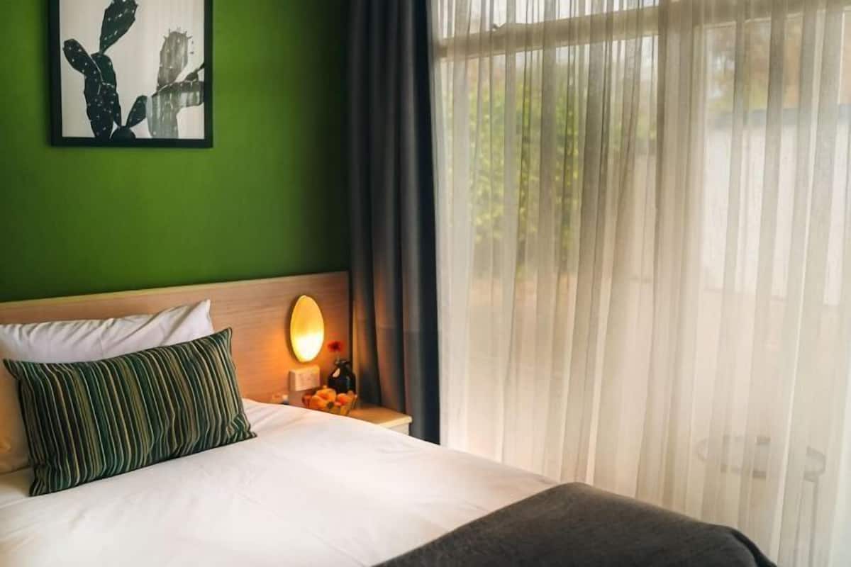 A hotel room with a bed, a nightstand with a lamp, a green wall, and a window with sheer curtains.