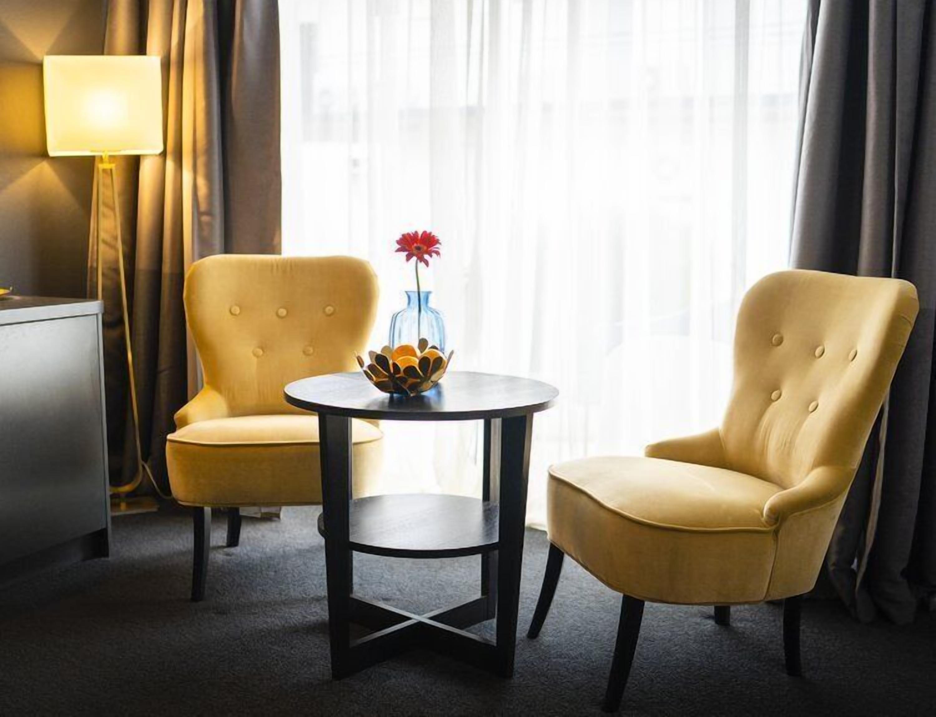 Una habitación de hotel con dos sillones amarillos, una mesita redonda con un jarrón decorativo y una lámpara.