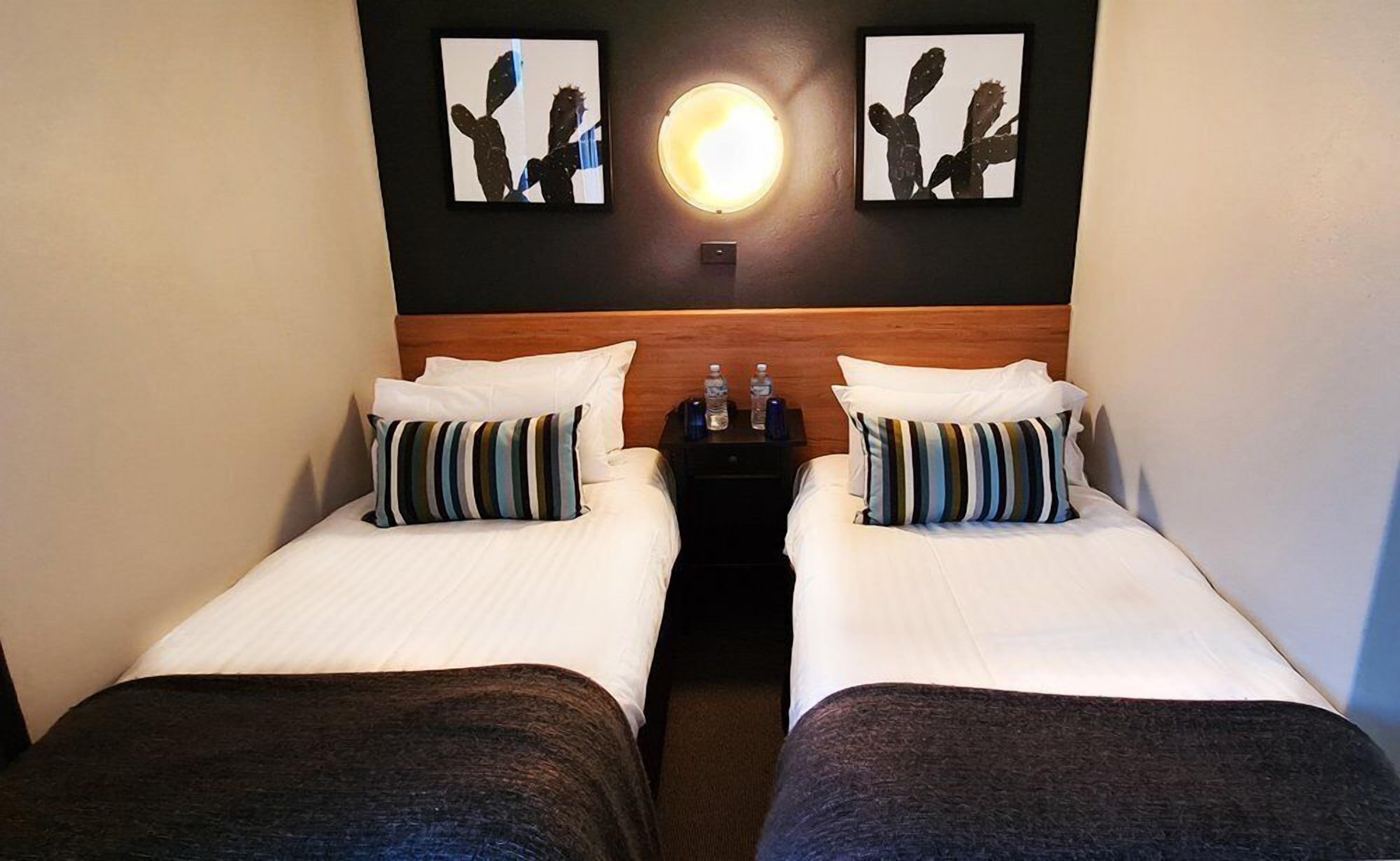 A hotel room with two single beds, each with a pillow and a blanket. There is a nightstand between the beds with two water bottles on it. Above the nightstand, there are two framed pictures of cacti.