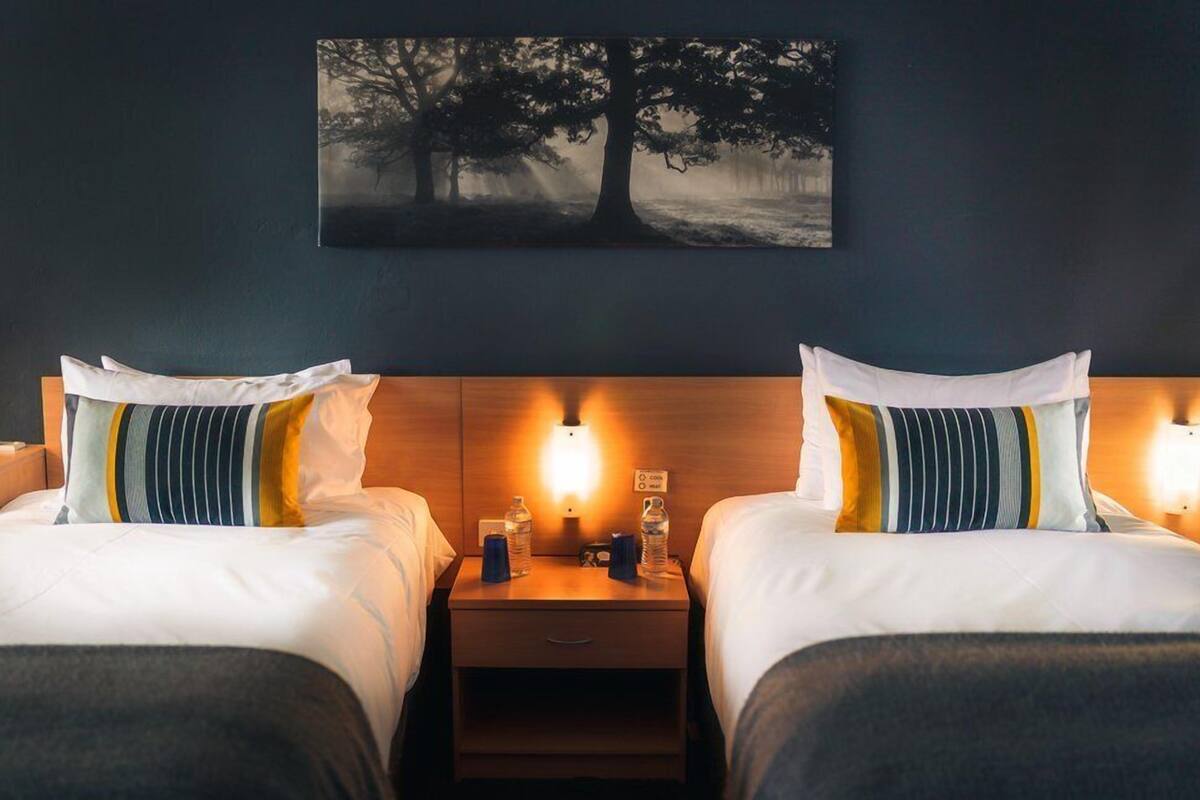 A hotel room with two beds, each with striped pillows, a nightstand with a lamp, and a painting of a tree on the wall.