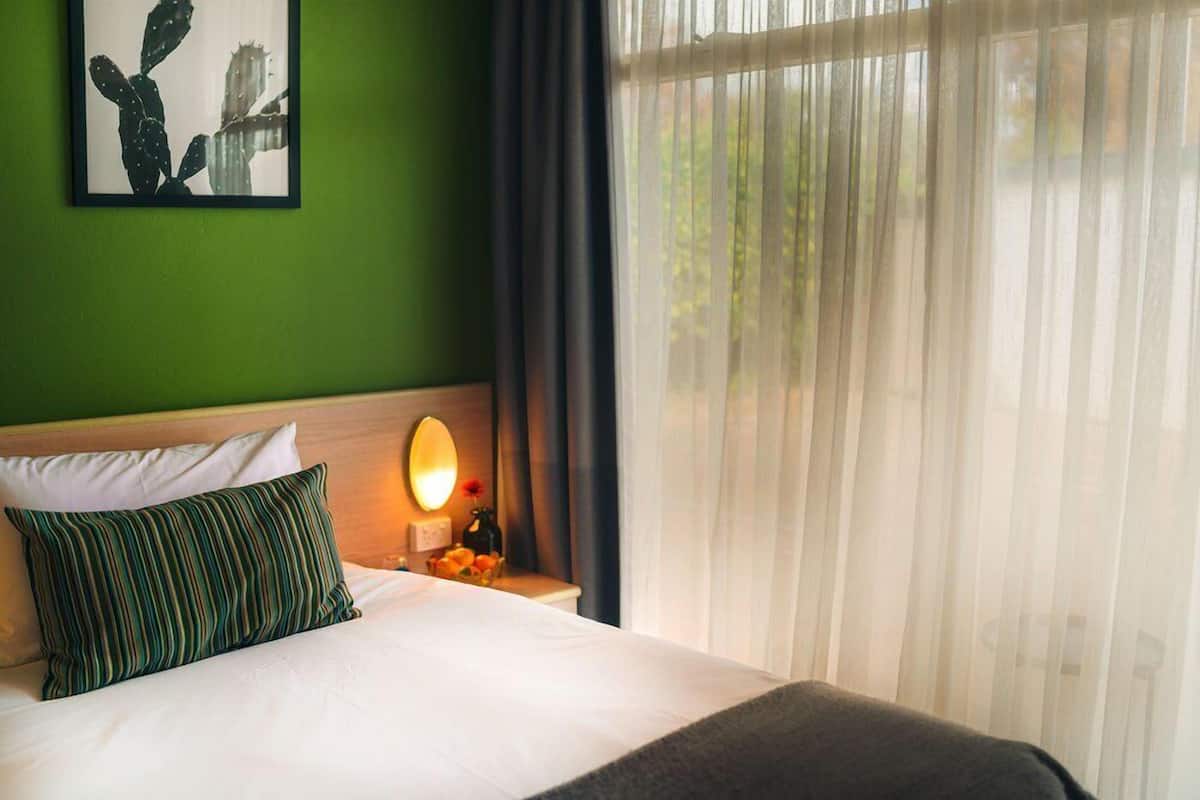 A hotel room with a bed, a nightstand with a lamp, a framed picture on the wall, and sheer curtains.