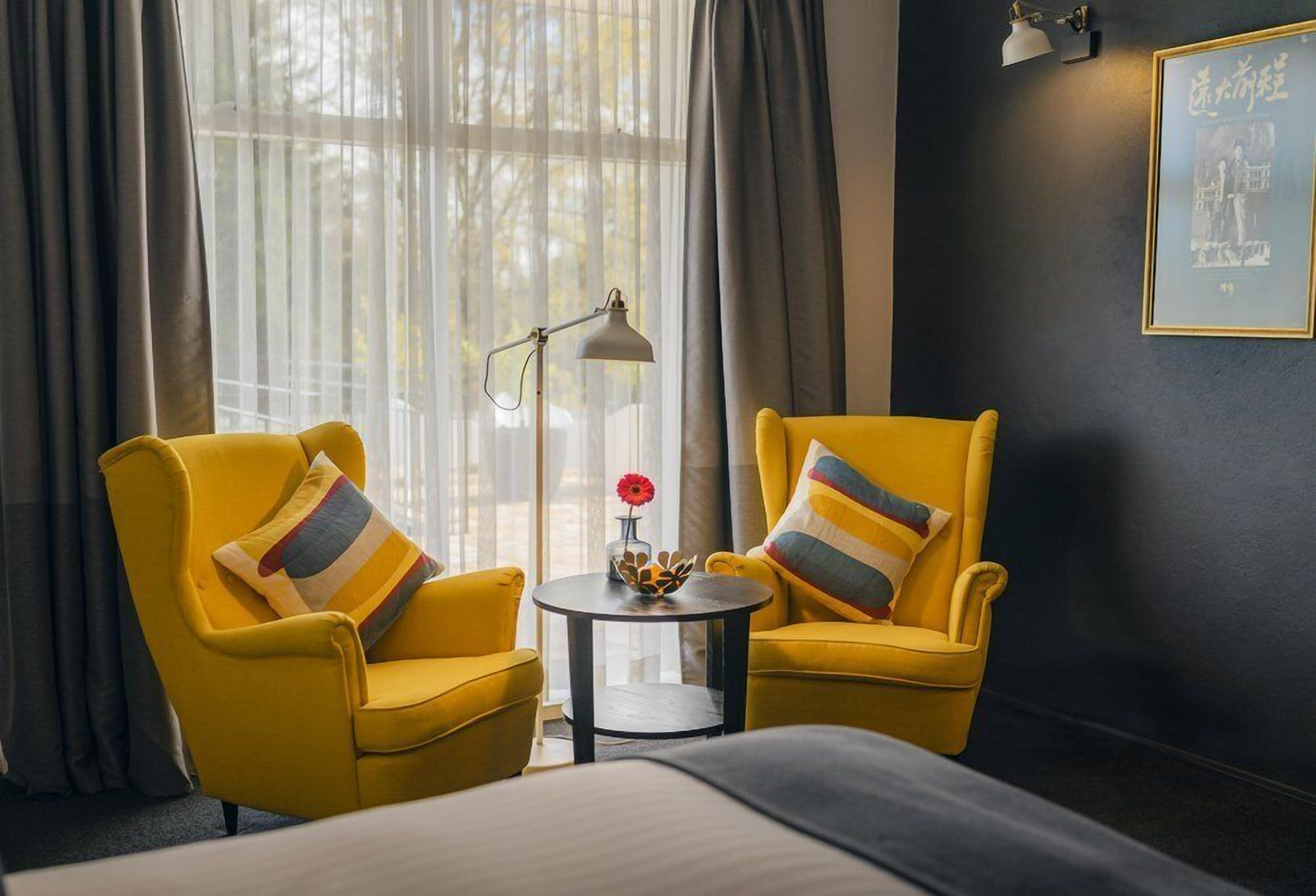 A hotel room with two yellow armchairs, a small round table with a vase and a red flower, and a bed with a white bedspread.