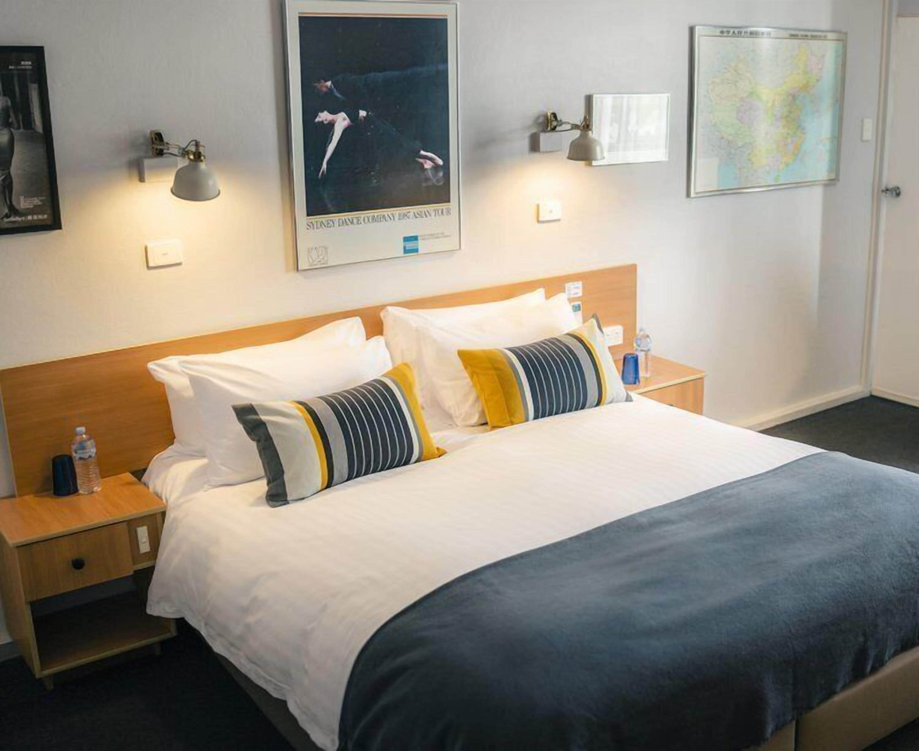 A hotel room with a large bed, two bedside tables, a wooden headboard, and wall-mounted artwork including a map and a dance poster.