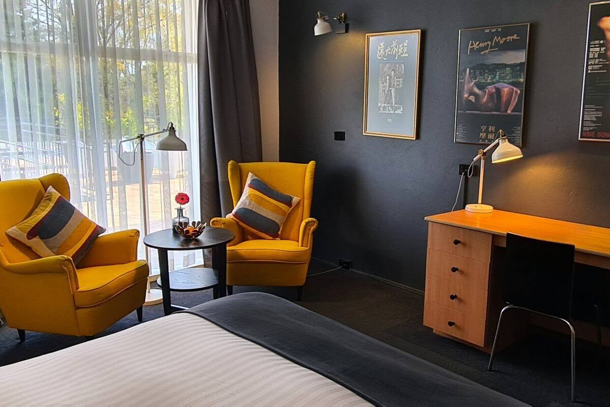 A hotel room with a bed, two yellow armchairs, a small round table, a desk with a chair, and a lamp.