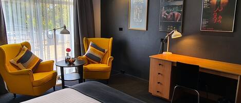 A hotel room with a bed, two yellow armchairs, a small round table, a desk with a chair, and a lamp.