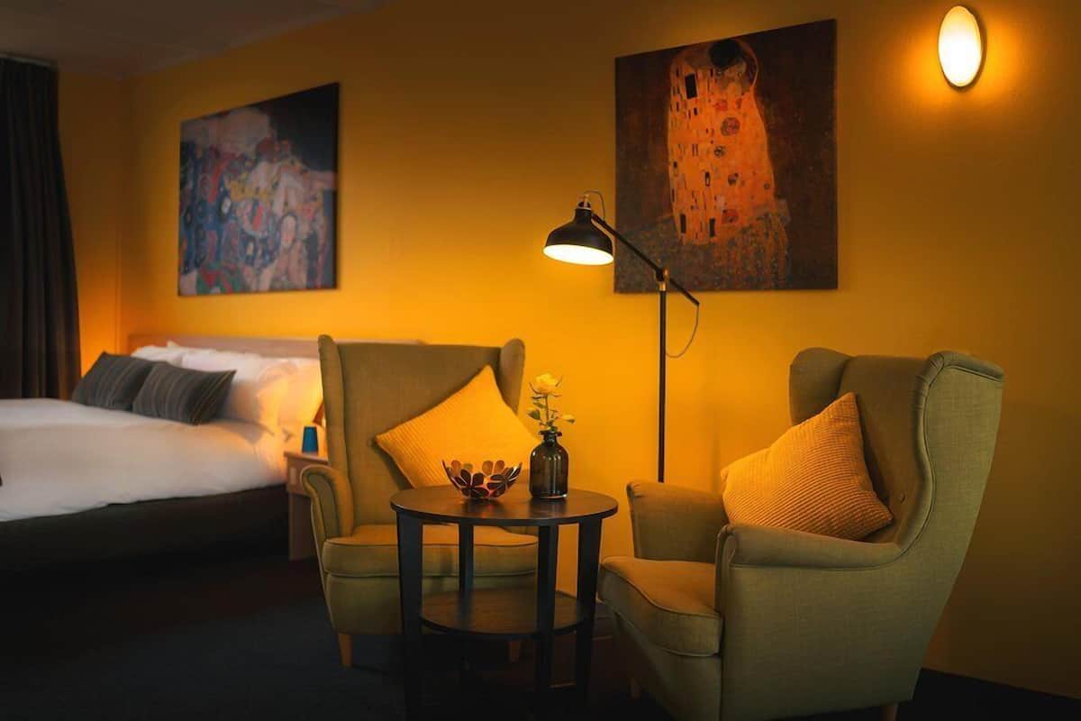 A hotel room with a bed, two armchairs, a small table with a vase, and two paintings on the wall.