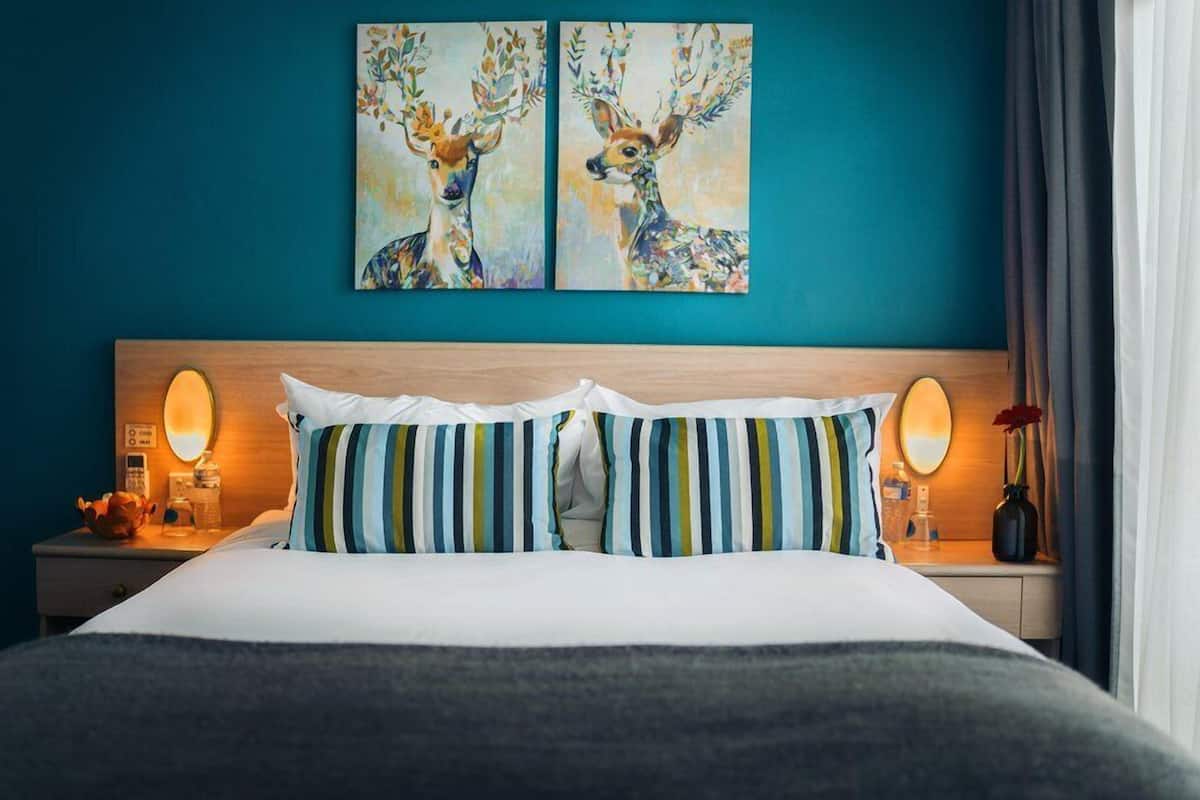 A hotel room with a teal wall, a wooden headboard, striped pillows, a bed with white bedding, two paintings of deer, bedside lamps, and a vase with flowers.