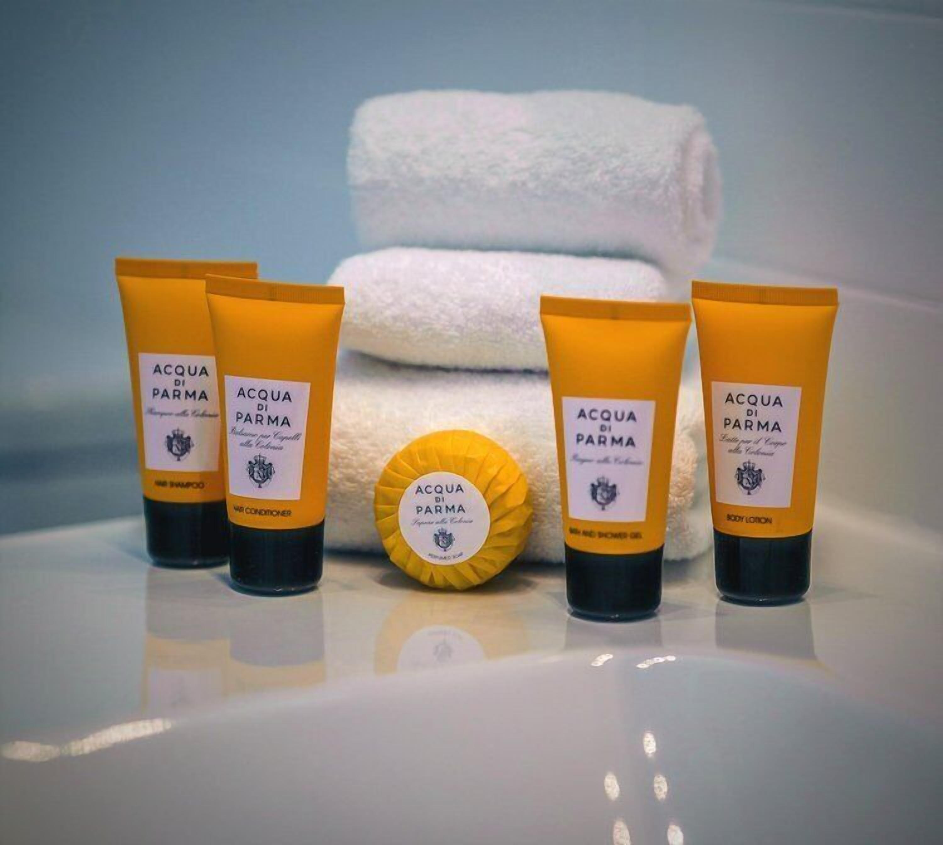 A set of toiletries including shampoo, conditioner, body lotion, and a soap bar, arranged on a reflective surface with a towel in the background.