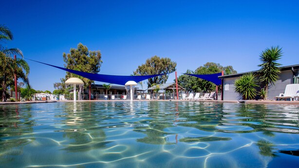 Una piscina cristalina con una vela de sombreo azul, sillas de descanso y un pabellón.