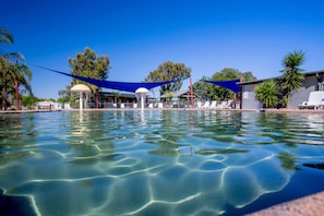 A clear swimming pool with a blue shade sail, lounge chairs, and a gazebo.
