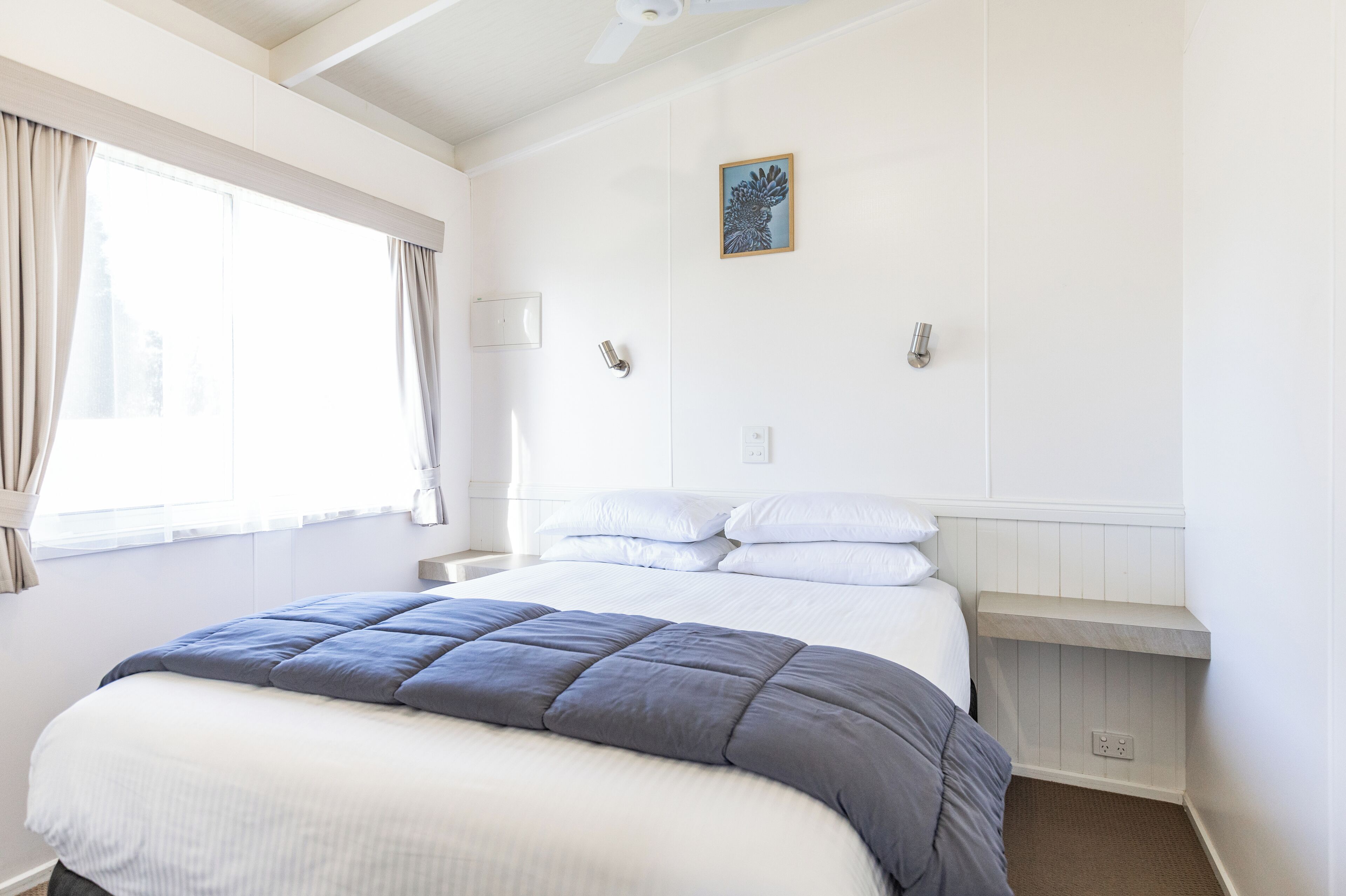 A neatly arranged bedroom with a large bed, a bedside table, a nightstand, a wall-mounted shelf, and a window with curtains.