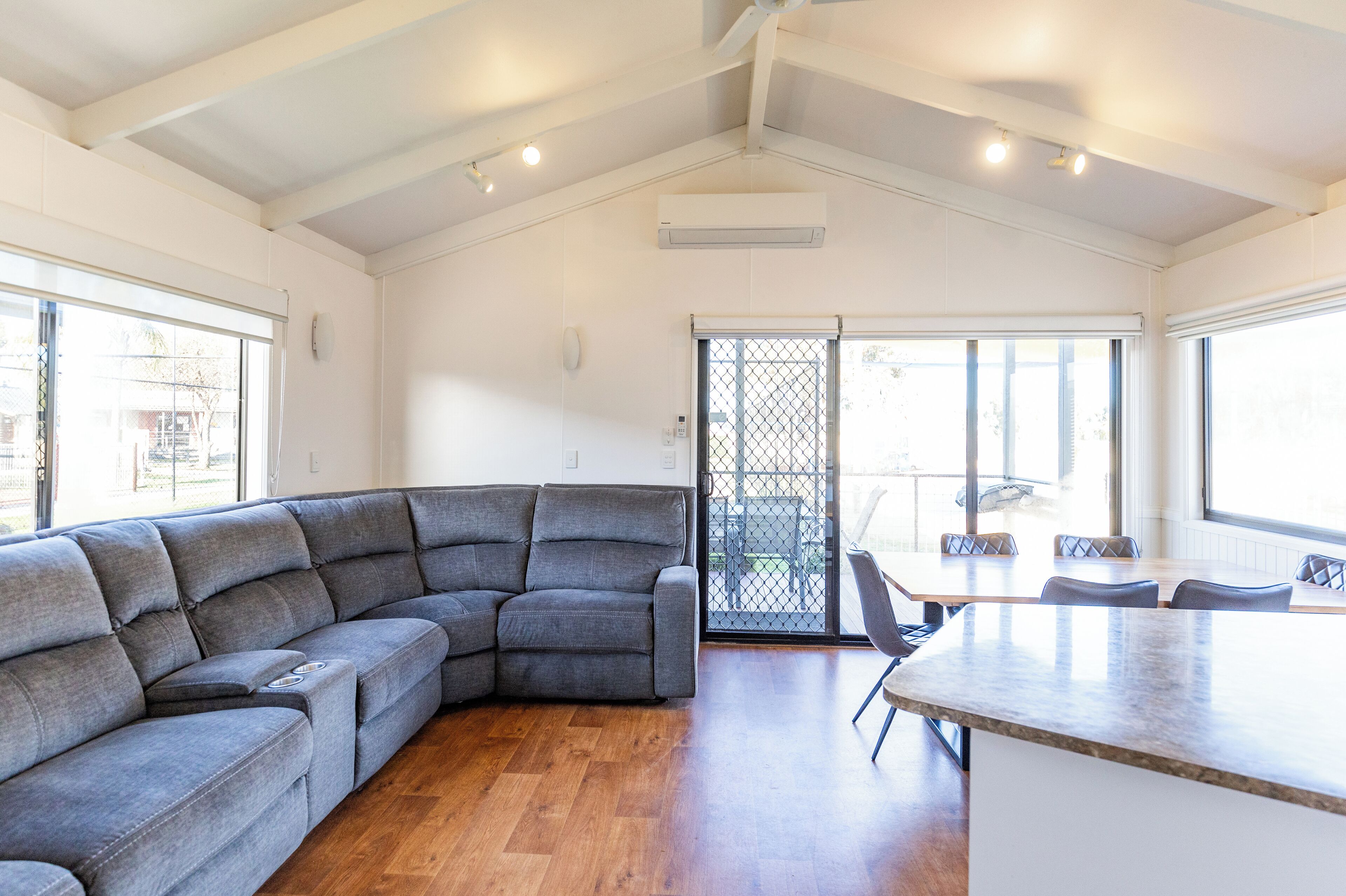 Un salon moderne avec un canapé d’angle, un coin repas comprenant une table et des chaises, et de grandes fenêtres qui laissent entrer la lumière naturelle.