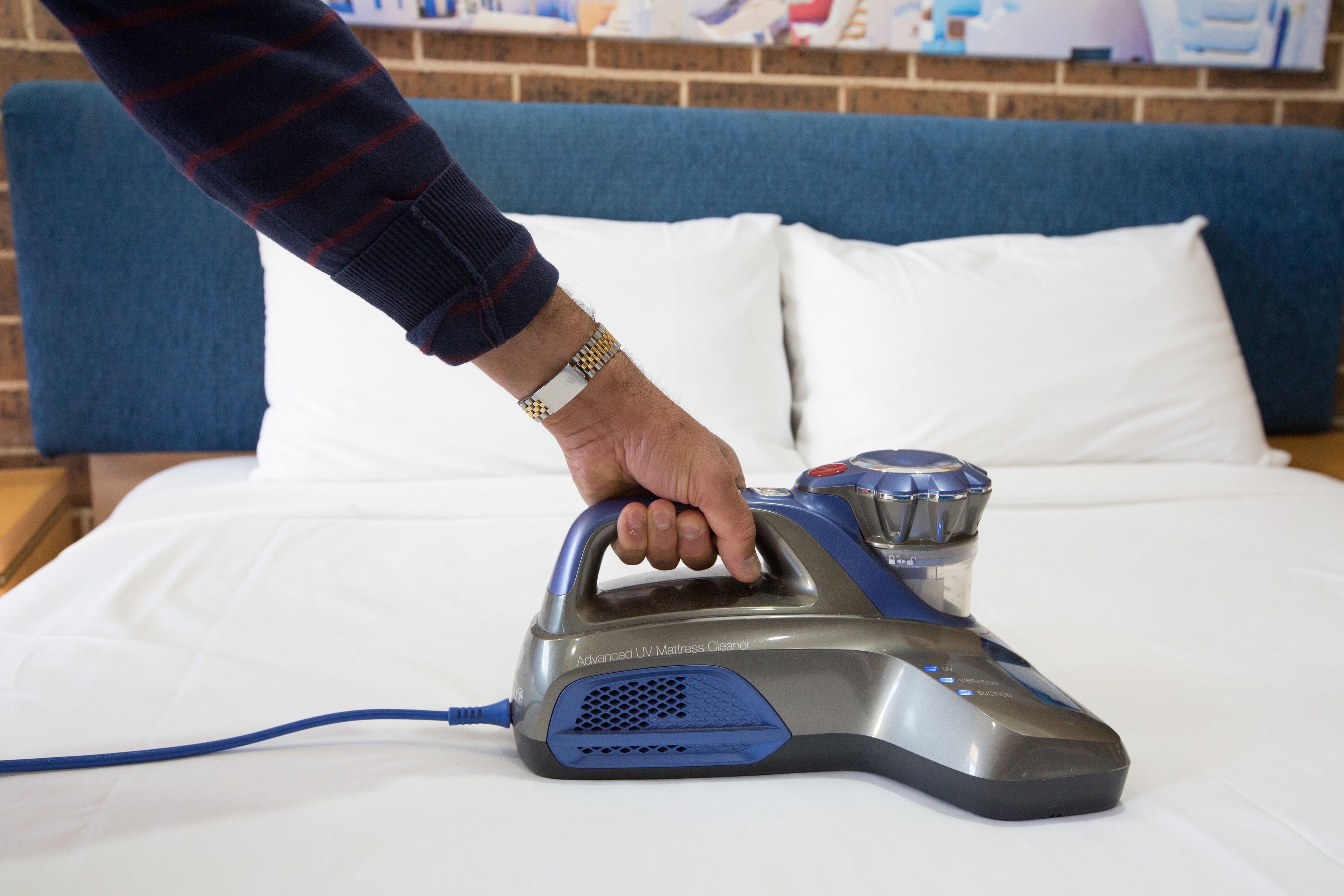 Una persona utilizando un limpiador UV avanzado para colchones sobre una cama con sábanas blancas y cabecera azul.