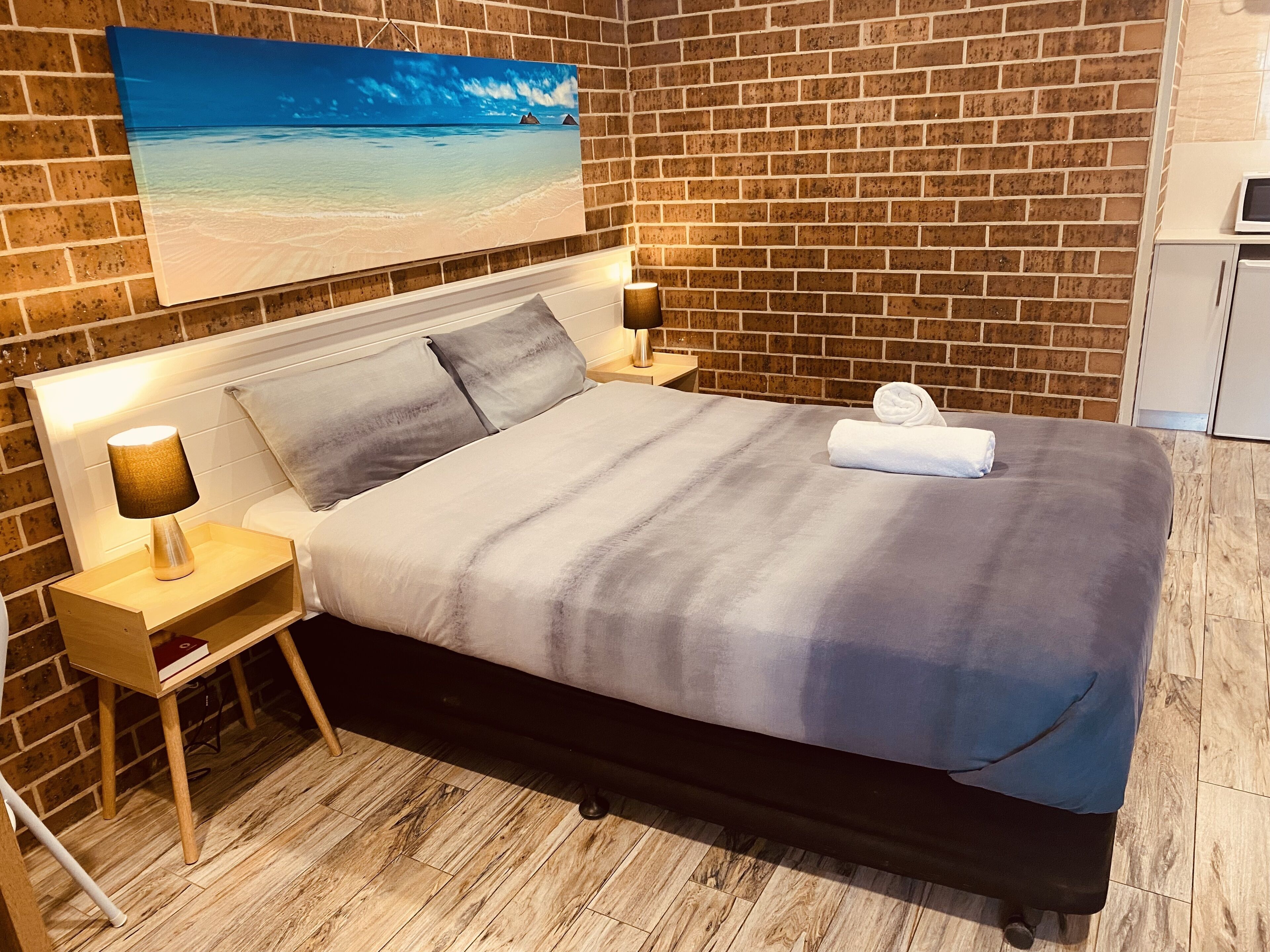 A hotel room with a brick wall, a bed with a grey and white bedspread, two bedside lamps, a wooden nightstand, and a painting of a beach scene above the bed.