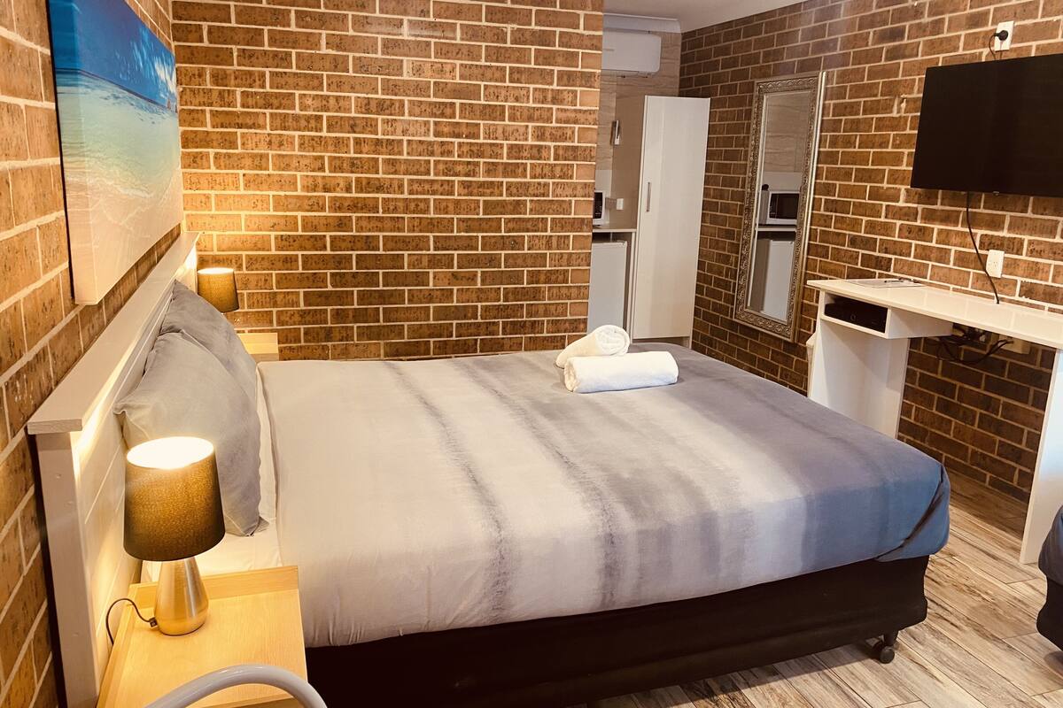 Hotellihuone, jossa on tiiliseinä, harmaalla peitteellä varustettu sänky, yöpöytälamppu, pyyhe, puinen yöpöytä, työpöytä, jossa on litteänäyttöinen televisio, ja peili seinällä.