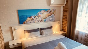 A hotel room with a bed, a nightstand, a wall-mounted air conditioner, a window with curtains, and a painting of a coastal village. - Buccaneer Motel Pet friendly (Long Jetty)