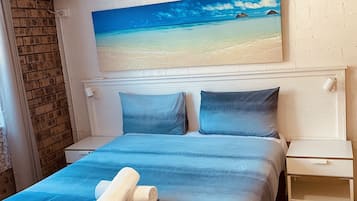 Una cama bien hecha con colcha y almohadas azules, una mesita de noche blanca y un cuadro con una playa en la pared.