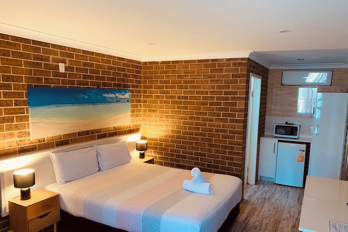 Hotellihuone, jossa on tiiliseinä, valkoisilla lakanoilla varustettu sänky, rantamaisemaa esittävä taulu, pieni keittokomero, jossa on mikroaaltouuni, ja puulattia.
