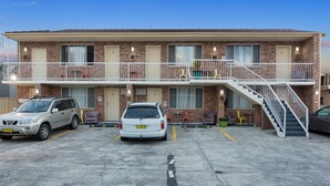 A two-story motel with a brick facade, a balcony on the upper floor, and a staircase leading to the entrance. There are two cars parked in front. - Buccaneer Motel Pet friendly (Long Jetty)