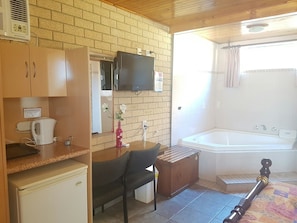A compact kitchen with a refrigerator, a microwave, and a coffee maker. There's a wooden cabinet, a small table with a chair, and a mirror. A television is mounted above the mirror. The room has a tiled floor and a wooden ceiling. - Lake Front Motel (The Entrance North)