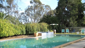 Una piscina con borde de azulejos, rodeada de césped y árboles. Hay sillas de descanso y una sombrilla grande cerca de la piscina.